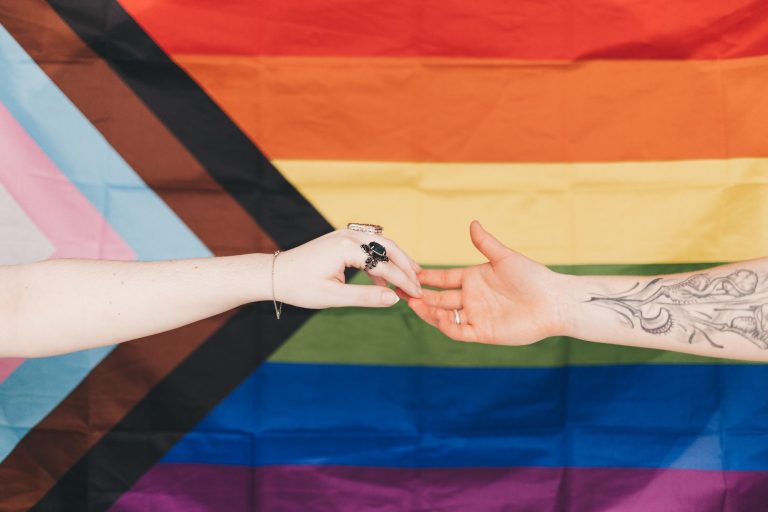 Two people's hands joined in front of a Progress Pride flag.