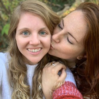 A photo of Edina cofounder Julia Sanders smiling into the camera while her partner, America Marvel, kisses her cheek